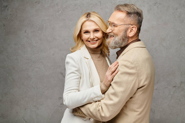 Een volwassen man en vrouw in elegante kleding omhelzen elkaar teder tegen een grijze achtergrond. - Foto, afbeelding