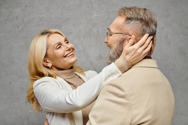 Un hombre y una mujer maduros en elegante atuendo abrazan amorosamente contra un fondo gris. - Foto, imagen