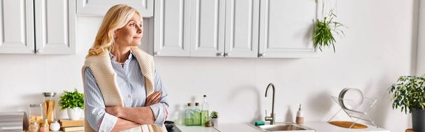 Μια ώριμη, ελκυστική γυναίκα ποζάρει δίπλα σε νεροχύτη κουζίνας στο σπίτι της, ακτινοβολώντας γαλήνια κομψότητα και χάρη.. - Φωτογραφία, εικόνα