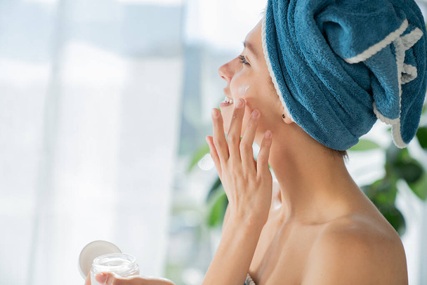 Atrakcyjna młoda kobieta nosząca ręcznik, nakładająca krem do twarzy na twarz jako rutynę pielęgnacyjną po prysznicu. Widok z boku szczęśliwy kobieta masowanie skóry twarzy i patrząc na okno. Pojęcie procedury pielęgnacji skóry. - Zdjęcie, obraz
