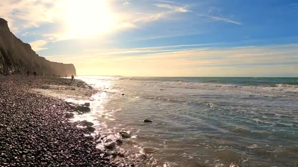 Questo filmato cattura la bellezza serena di una spiaggia di ciottoli illuminata dal sole, con maestose scogliere che sovrastano le onde che si infrangono dolcemente. La luce dorata del sole riflette l'acqua, creando uno scintillante - Filmati, video