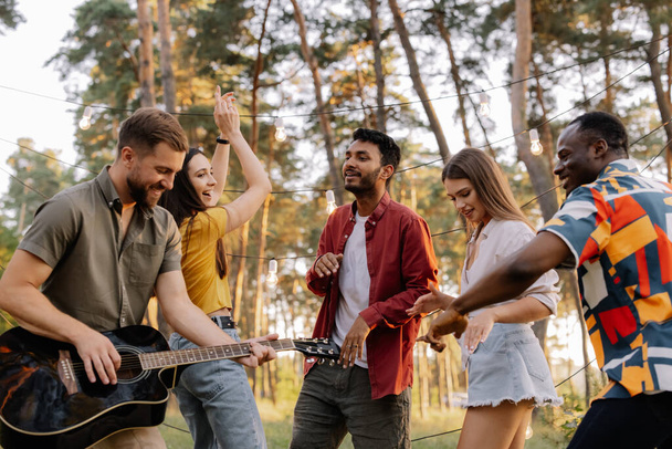 Grupo multirracial de pessoas, homem hipster barbudo tocando guitarra e amigos dançando, cantando e se divertindo - Foto, Imagem