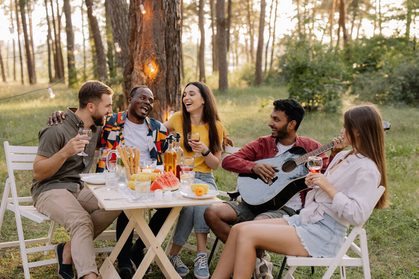 Spotkanie wielorasowej grupy przyjaciół grających na gitarze, śpiewających, jedzących kolację i pijących wino podczas imprezy w lesie - Zdjęcie, obraz