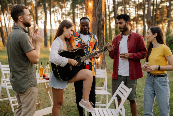 Atrakcyjna hipsterka grająca na gitarze wygrywająca z mężczyznami na kolacji z wielorasowymi przyjaciółmi - Zdjęcie, obraz