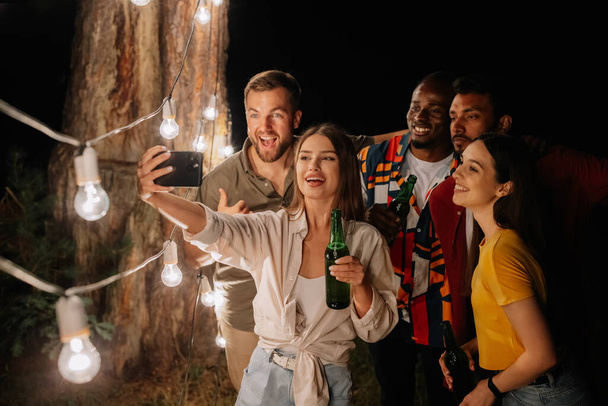 Un grupo de amigos multirraciales divirtiéndose y bebiendo cerveza se toman selfies cerca de lámparas colgantes por la noche - Foto, Imagen