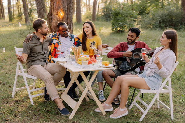Ontmoeting van multiraciale vrienden die gitaar spelen, zingen, eten en wijn drinken tijdens een feest in het bos - Foto, afbeelding