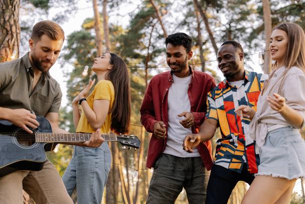 Multiraciale groep mensen, bebaarde hipster man spelen gitaar en vrienden dansen, zingen en plezier hebben - Foto, afbeelding