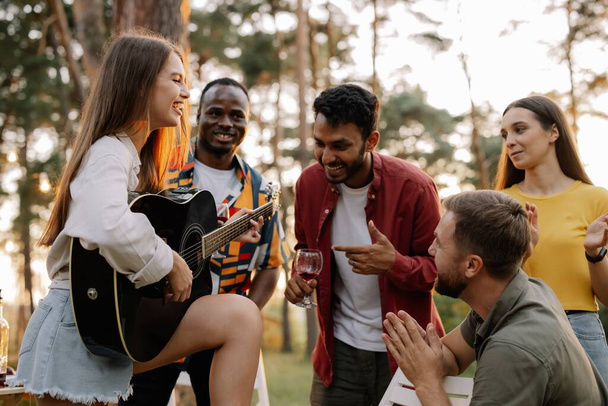 Attractive hipster woman playing guitar winning over men at dinner party with multiracial friends - Photo, Image