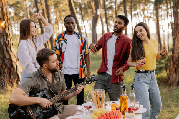 Wielokulturowa grupa ludzi, brodaty hipster człowiek gra na gitarze i przyjaciele tańczą i dobrze się bawią - Zdjęcie, obraz
