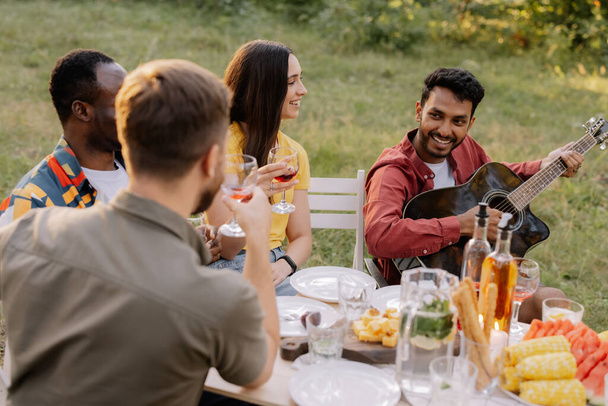 Multiracial group of people, Indian hipster man playing guitar and friends eating dinner and drinking wine during party in the forest - Photo, Image