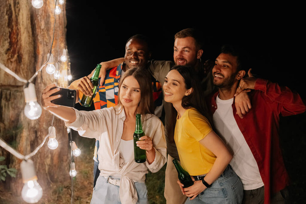 Um grupo de amigos multirraciais se divertindo e bebendo cerveja tirar selfies perto de lâmpadas penduradas à noite - Foto, Imagem