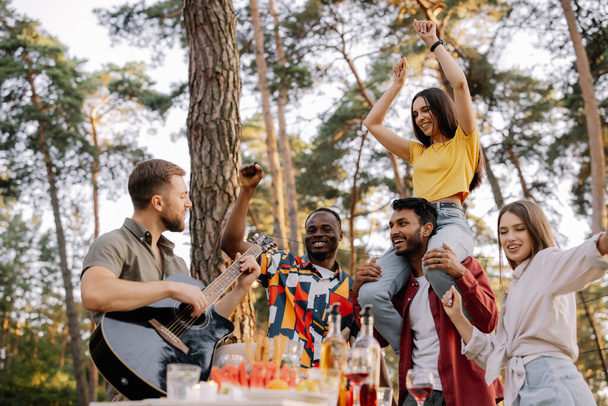 Grupo multicultural de pessoas, homem hipster barbudo tocando guitarra e amigos dançando, cantando e se divertindo - Foto, Imagem