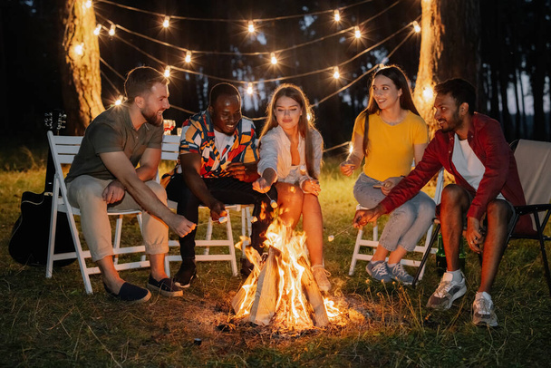 Skupina multiraciálních klidných přátel pečení marshmallows zatímco sedí u táboráku - Fotografie, Obrázek