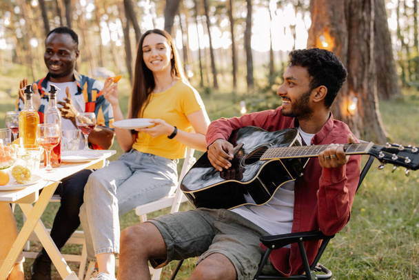 Wielorasowa grupa ludzi, indyjski hipster gra na gitarze i przyjaciele jedzą obiad i piją wino podczas imprezy w lesie - Zdjęcie, obraz