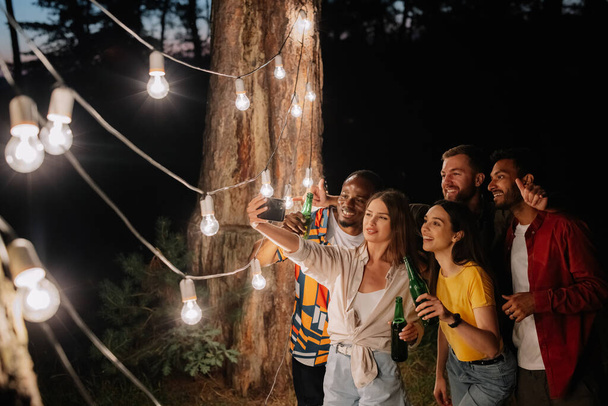 Un grupo de amigos multirraciales divirtiéndose y bebiendo cerveza se toman selfies cerca de lámparas colgantes por la noche - Foto, imagen