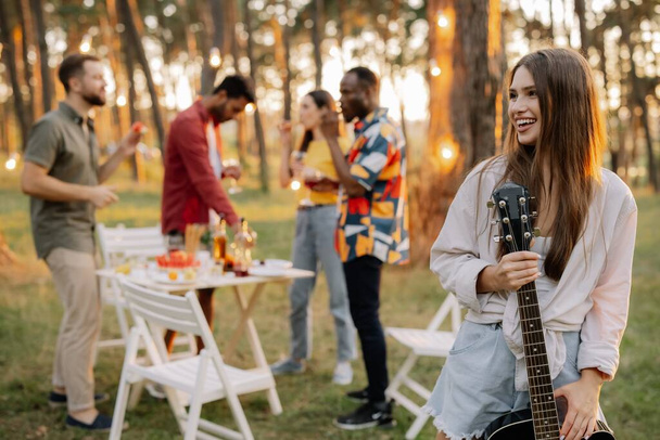 Красивая хипстерша, играющая на гитаре на фоне званого ужина у многорасовых друзей - Фото, изображение