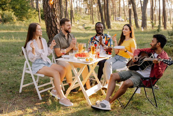 森でパーティー中にギターを弾き,歌い,夕食を食べ,ワインを飲む友人の多種多様なグループの会合 - 写真・画像