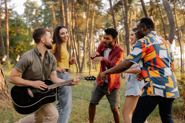 Мультирасовая группа людей, бородатый хипстер, играющий на гитаре, и друзья танцуют, поют и веселятся - Фото, изображение