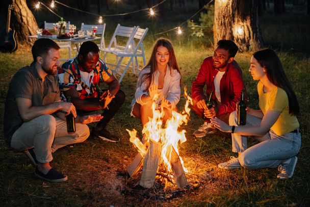Mnohostranní přátelé sedí u ohně, popíjejí pivo a baví se na večírku - Fotografie, Obrázek