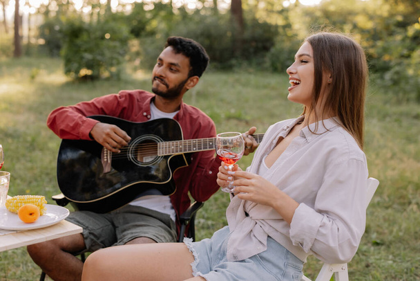 Multiraciální skupina lidí, indický hipster muž hraje na kytaru a přátelé jíst večeři a pít víno během párty v lese - Fotografie, Obrázek