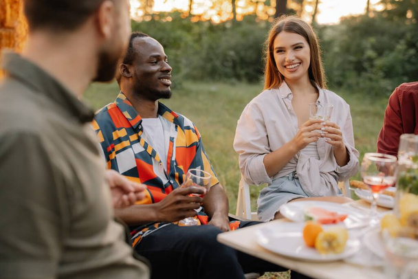 Gruppo multirazziale di amici felici seduti intorno al tavolo a bere vino parlando a cena nella foresta estiva serale - Foto, immagini