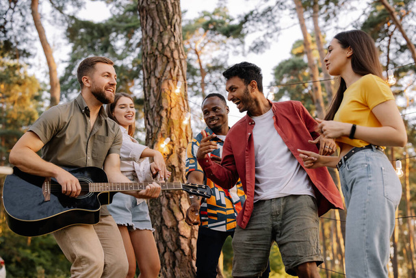 Мультирасовая группа людей, бородатый хипстер, играющий на гитаре, и друзья танцуют, поют и веселятся - Фото, изображение