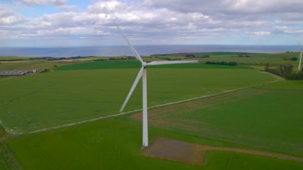 AERIAL: Rotierende Windmühlen inmitten grüner Felder an der windigen schottischen Küste. Hohe und majestätische moderne Strukturen, die natürliche Elemente für eine nachhaltige Stromerzeugung nutzen. - Filmmaterial, Video