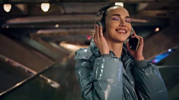 Vidám boldog tizenéves lány zenét hallgat vezeték nélküli headset tartja a modern okostelefon közeli. Mosolygó nyugodt nő táncol tapintatos energikus dal eszik nyalóka. Fényes barna élvezze az éjszakai életet - Fotó, kép