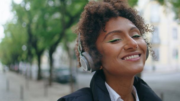 Carefree αφροαμερικανή κοπέλα ακούγοντας μουσική από ασύρματα ακουστικά στο δρόμο από κοντά. Χαρούμενη χαμογελαστή γυναίκα που κοιτάζει την κάμερα φορώντας μοντέρνα ακουστικά σε εξωτερικούς χώρους. Η όμορφη κυρία απολαμβάνει τον όμορφο ήχο. - Φωτογραφία, εικόνα