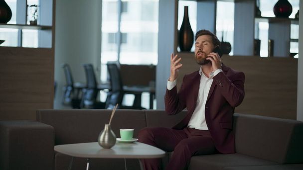 Angry realtor talking smartphone in modern comfortable office interior. Stressed beard man professional making call on sofa. Focused manager discussing plans near big window. Corporate person concept - Photo, Image