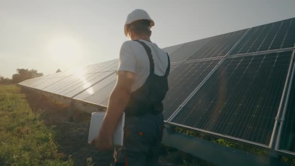 Un trabajador masculino con documentación realiza tareas de trabajo en una estación de paneles solares. Supervisión del funcionamiento del sistema de generación de electricidad. Concepto de electricidad verde. Imágenes 4K de alta calidad - Metraje, vídeo