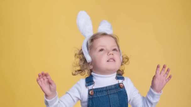 Un bambino felice che indossa orecchie da coniglio e tute si alza su uno sfondo giallo, sorridendo e gesticolando con il pollice, divertendosi e condividendo la gioia con un dito giocoso. - Filmati, video