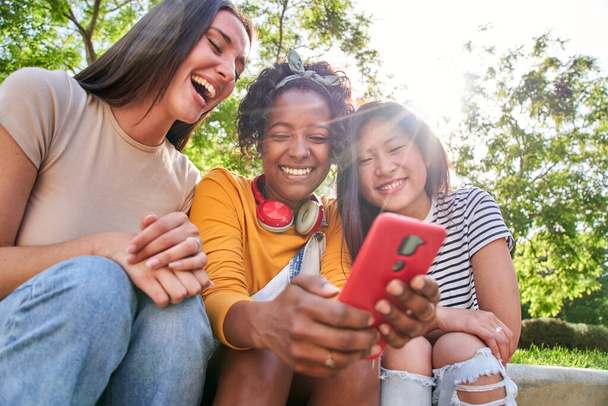 Grupo amigos alegres utilizando móvil sentado al aire libre. Tres mujeres multirraciales riendo viendo las redes sociales en el teléfono inteligente. Gen z chicas divirtiéndose viendo algo en el dispositivo. Jóvenes juntos - Foto, imagen