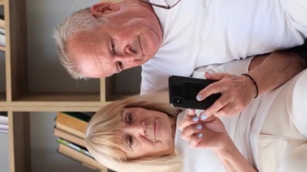 O casal idoso compartilha alegremente um sorriso enquanto se senta em um sofá, olhando para um telefone celular enquanto gentilmente tocam o cabelo uns dos outros e trocam gestos.. - Filmagem, Vídeo