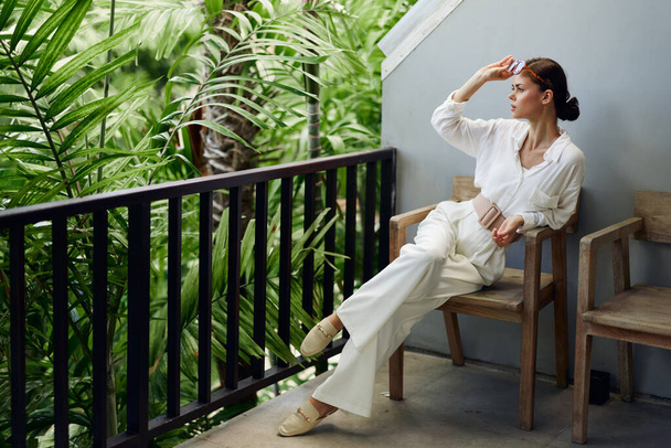 Uśmiechnięta kobieta ciesząca się tropikalnym wypoczynkiem na słonecznym balkonie z widokiem na spokojny ocean Ona relaksuje się w modnym stroju, nosi okulary przeciwsłoneczne i popijając orzeźwiającego drinka Piękna przyroda - Zdjęcie, obraz