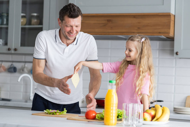 A friendly family from smiling father and daughter stand in the kitchen in a joyful mood, have fun, hug and laugh together. A young man dad and little girl child demonstrate positive emotions - Photo, Image