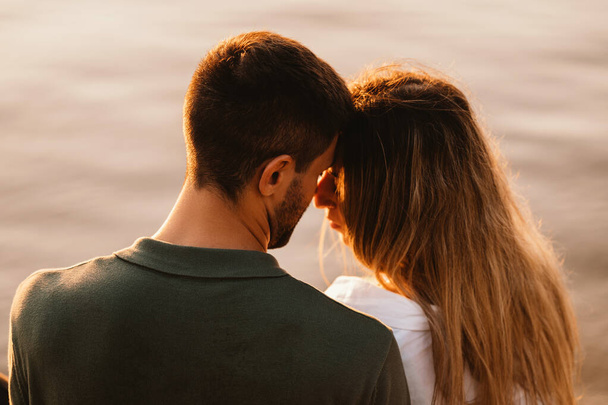 close-up schot van een man en een vrouw aanraken van hun voorhoofd bij zonsondergang whie zitten in de buurt van een meer. - Foto, afbeelding