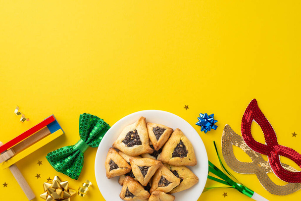 Top view shot of Purim festivity essentials, beleértve a töltött zsebű cookie-k tányéron, szalagcsillagok, álarcos maszkok, nyakkendő, zajkeltő, rendezett élénk sárga háttér üres terület szöveges - Fotó, kép