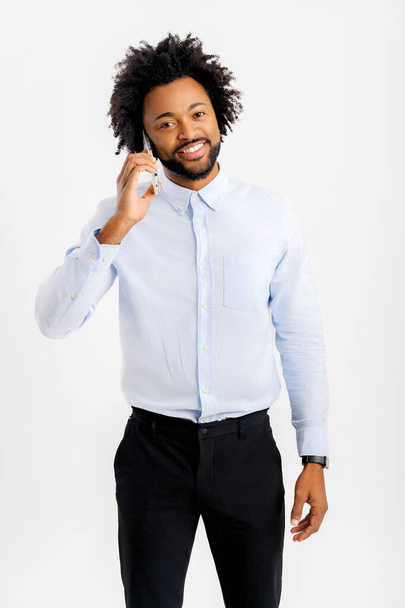 Alegre hombre afroamericano en ropa formal hablando en el teléfono inteligente aislado en blanco, empleado de oficina masculino tener una agradable conversación telefónica, utilizando el nuevo móvil de moda, mira a la cámara - Foto, imagen