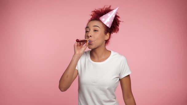 Щаслива афро-американська руда кучерява жінка в день народження вибухає свисток святкуючи свято. Радісна усміхнена жінка в білій футболці на ізольованому рожевому тлі з копіювальним простором - Кадри, відео