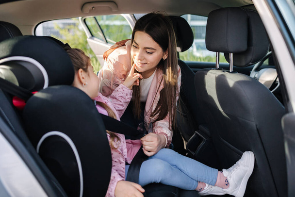 Atractiva madre joven puso a su hija en un asiento de coche y se abrocha los cinturones de seguridad. Las mujeres se preocupan por los niños. Protección durante el viaje en el coche. Foto de alta calidad - Foto, imagen
