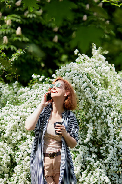 Červenovlasá žena mluví po telefonu. Příroda volá na volné noze život radostný pracovní okamžik pod kvetoucími větvemi - Fotografie, Obrázek
