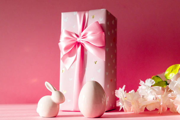 Figurines en porcelaine d'œufs de Pâques, un lapin, un grand coffret cadeau, et une branche de fleurs de pommier se tiennent sur un fond rose, vue latérale rapprochée avec mise au point sélective. - Photo, image