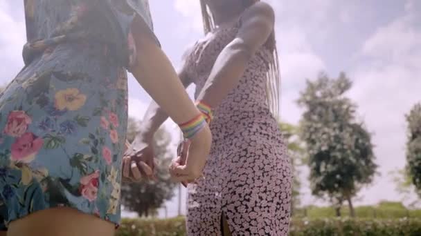 Close-up van achteren van een onherkenbaar multi-raciaal lesbisch koppel dat vreedzaam hand in hand door een parktuin loopt. Twee jonge geliefden met regenboogvlag armbanden. Homoseksuele betrekkingen. - Video