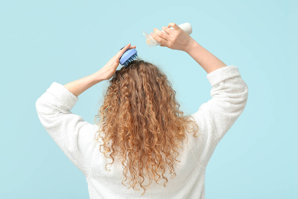 Young woman using hair scalp massager with bottle of shampoo on blue background, back view - Photo, Image