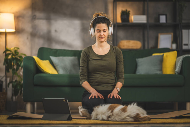 Yetişkin bir kadın Kafkas kadın milenyumu kulaklıkları online rehberli meditasyon için kullanıyor evde gözleri kapalı farkındalık yogası yapıyor gerçek insanların kişisel bakım kavramını taklit ediyor. - Fotoğraf, Görsel