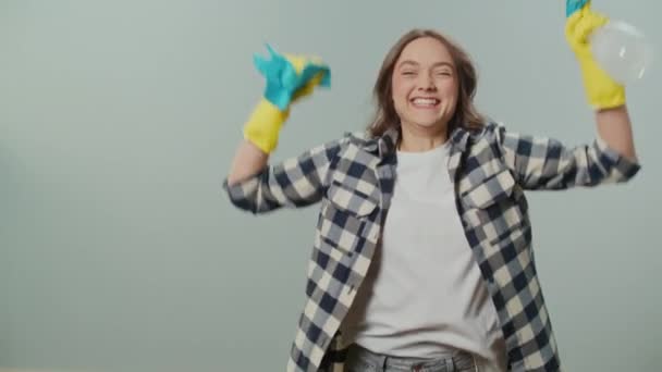 Een portret van een gelukkige jonge vrouw in gele handschoenen, het vasthouden van een reinigingsspray fles en doek, Verheugt zich over het succes in het schoonmaken, Springt voor vreugde op de grijze achtergrond.Decluttering Hacks. - Video