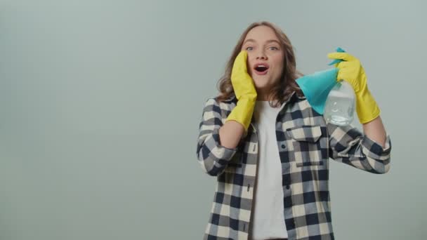 Retrato de uma jovem mulher surpreso em luvas amarelas, segurando uma garrafa de spray de limpeza e Rag no fundo cinzento.Uma dona de casa feminina está se preparando para limpar. Ferramentas de limpeza multifuncionais. - Filmagem, Vídeo