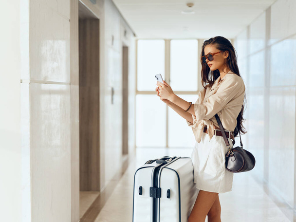 Delicia de los viajeros: Hermosa joven esperando su vuelo, sosteniendo un teléfono inteligente y una maleta, abrazando el espíritu navideño en una terminal del aeropuerto - Foto, imagen