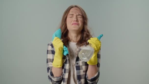Sucio molesto enojado mujer joven ama de casa en guantes amarillos sosteniendo una botella de aerosol de limpieza y trapo, casarse en el fondo gris. Consultas virtuales de limpieza. - Metraje, vídeo